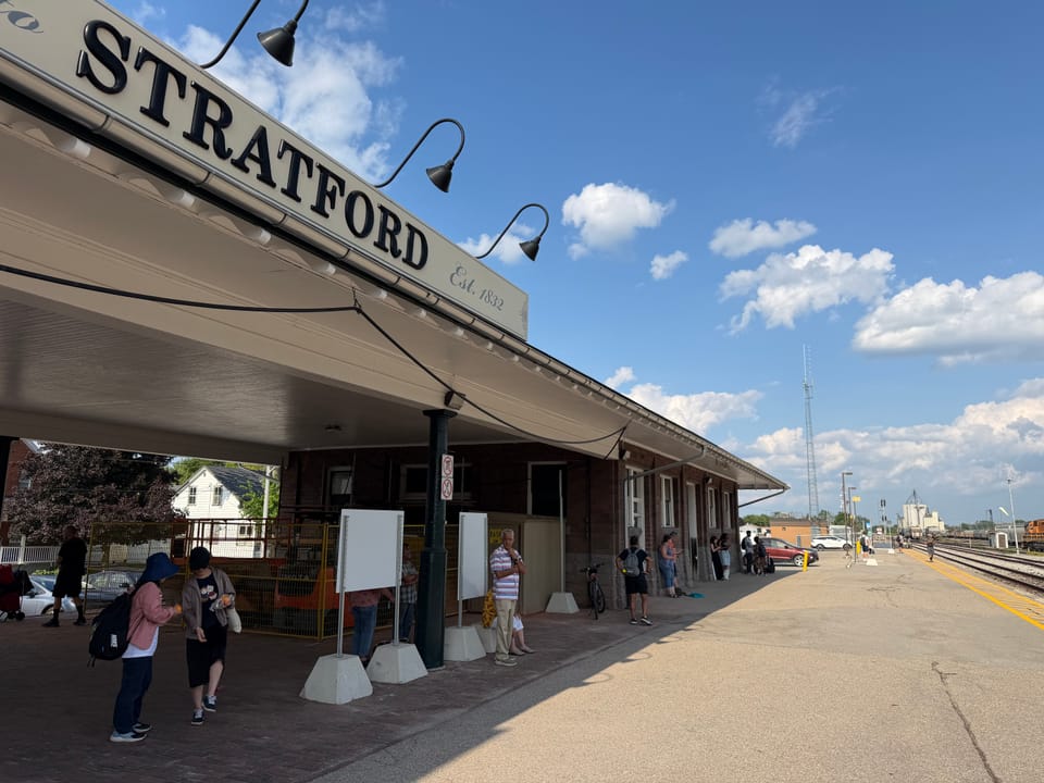 People waiting in the shade of a train station building with a big sign saying Stratford Est. 1832